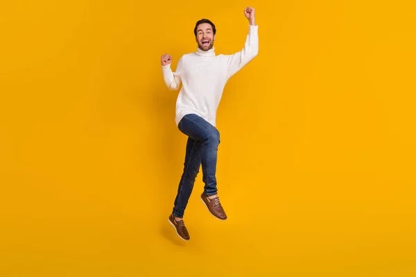 Photo of lucky pretty guy wear white pullover jumping high rising fists isolated yellow color background.