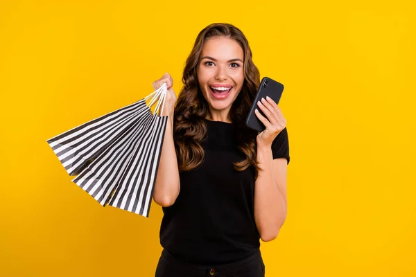 Photo of charming excited young lady dressed black clothes holding shopping bags modern device smiling isolated yellow color background.