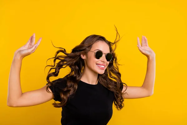 Photo of charming excited young lady dressed black clothes dark eyewear wind blowing smiling isolated yellow color background.