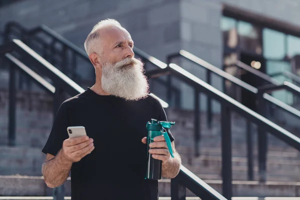 Photo of old beard sportive man run drink water hold telephone wear black t-shirt outside in city.
