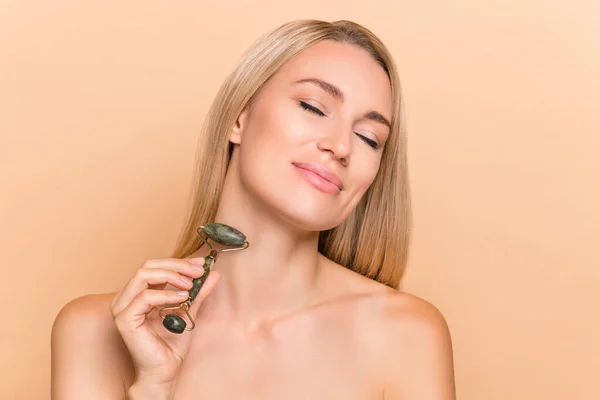 Photo of lady use jade stone massager enjoy aesthetic wellbeing peeling effect isolated over beige pastel color background.