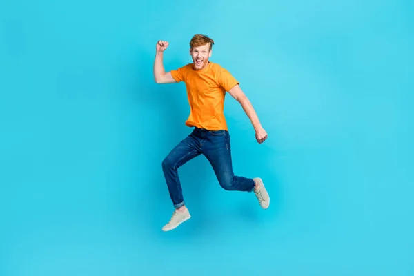 Full length photo of hooray ginger hair guy jump wear t-shirt jeans sneakers isolated on blue color background.