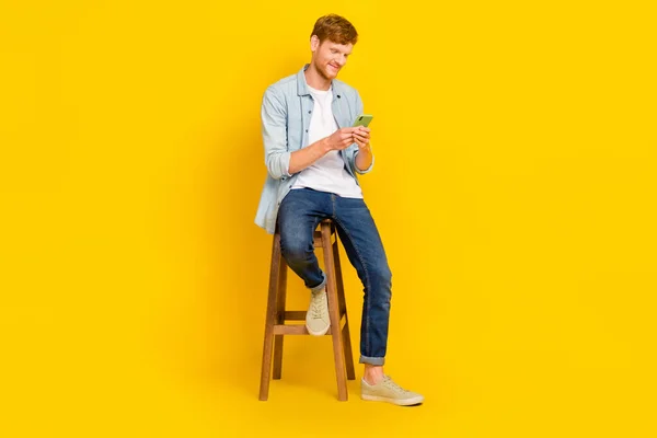 正の魅力的な男の写真を着てデニムシャツチャット現代的なデバイス座って椅子隔離された黄色の背景 — ストック写真