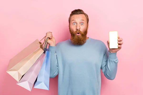 ピンク色の背景に隔離されたショッピングバッグスマートフォンを保持している青のスウェットシャツを身に着けている生姜の髪型と面白いファンキーな男の写真 — ストック写真