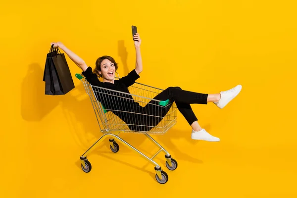 かわいい興奮女性の写真黒シャツ上昇腕に乗るショッピングトレイ上昇デバイスバッグ隔離された黄色の色の背景 — ストック写真