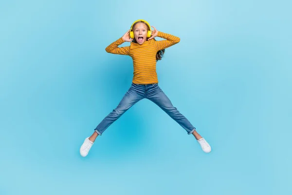 Full size photo of excited girl dressed yellow pullover denim pants flying jumping impressed playlist isolated on blue color background.