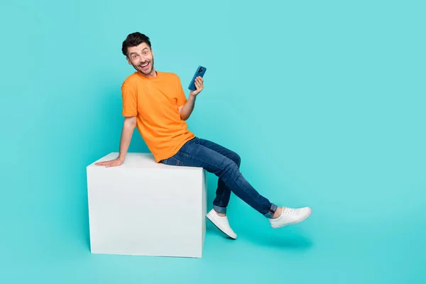Full body profile side photo of excited guy sit white cube using gadget like posts isolated on cyan color background.