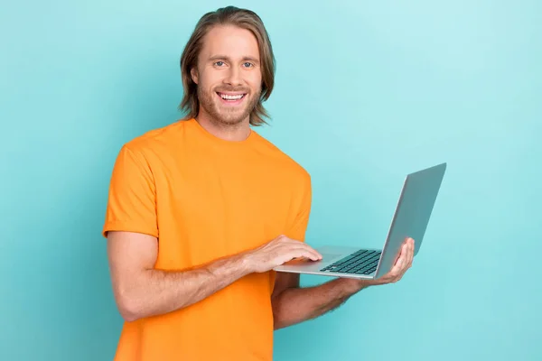 若いひげを生やした魅力的なハンサムな男の写真は オレンジのTシャツの作業ノートパソコンを身に着けているそれはプログラマーのおもちゃの笑顔は青の色の背景に隔離された — ストック写真