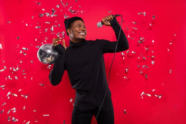 Portrait of attractive cheerful guy singing hit decor flying rest holding disco ball isolated over bright red color background.