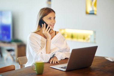 Beyaz tişörtlü güzel bir kadın evinde laptopuyla ve masada bir fincan kahveyle bir telefon görüşmesi yapıyor..
