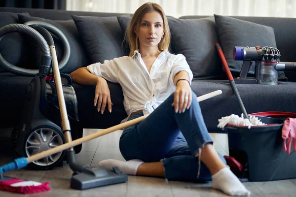Woman sitting on the floor near sofa with many cleaning tools