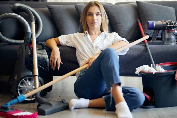 Kanepenin yanında yerde oturan bir sürü temizlik malzemesi olan kadın.