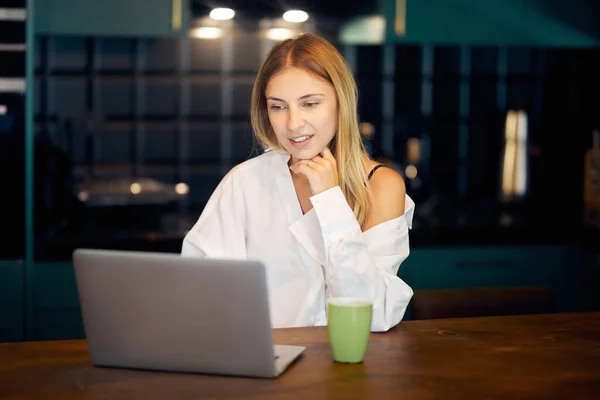 Evde çalışan beyaz gömlekli genç bir kadın. Masasında bir fincan kahveyle dizüstü bilgisayar kullanıyor.