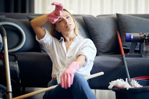 Tired after cleaning woman sitting on the floor near sofa with many cleaning tools