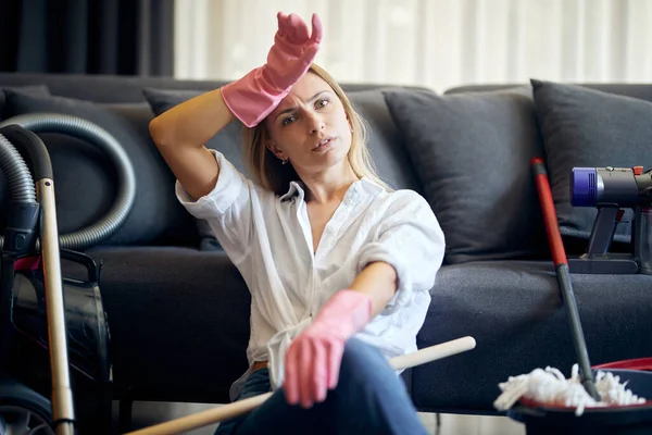 Tired after cleaning woman sits on the floor near sofa with many cleaning tools