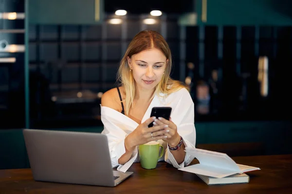 Beyaz tişörtlü genç sarışın kadın mutfaktaki defterin yanında cep telefonu kullanıyor.