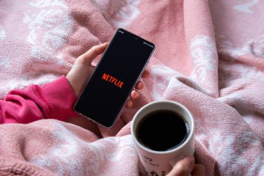 Afyonkarahisar, Türkiye - 9 Şubat 2022: Netflix logolu cep telefonu tutan kadın. Netflix popüler bir video yayın platformudur. Kadın yatakta oturuyor, çay ya da kahve içiyor ve film seyrediyor..