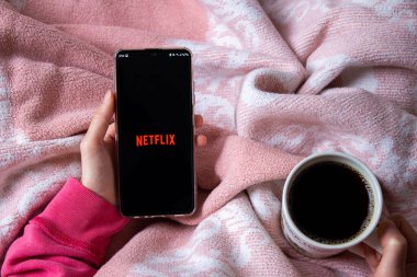 Afyonkarahisar, Türkiye - 9 Şubat 2022: Netflix logolu cep telefonu tutan kadın. Netflix popüler bir video yayın platformudur. Kadın yatakta oturuyor, çay ya da kahve içiyor ve film seyrediyor..