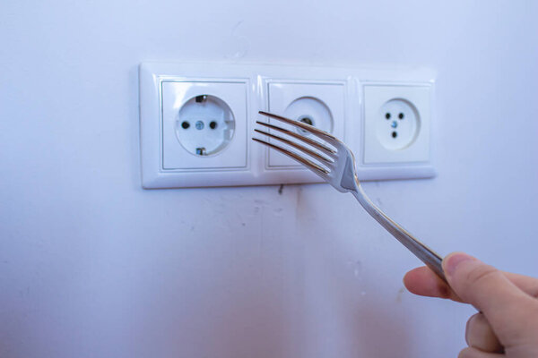 Children is approaching the electrical outlet with a fork in her hand. Concept of hazard for children at home.