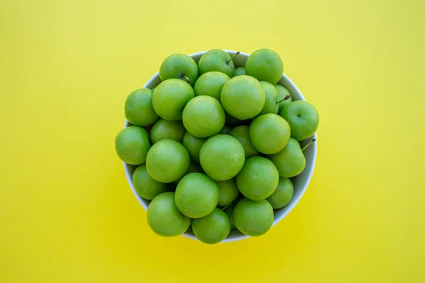 Green plums in a bowl isolated on yellow background. Top view photo of green plum fruits.
