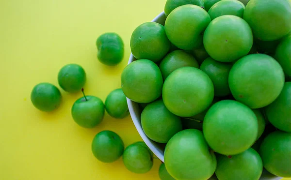 Green plums in a bowl on yellow background. Top view photo of plum fruits.