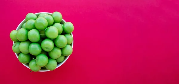 Green plums in bowl isolated on red background. Top view photo of sour plum fruits with space for text. High quality photo.