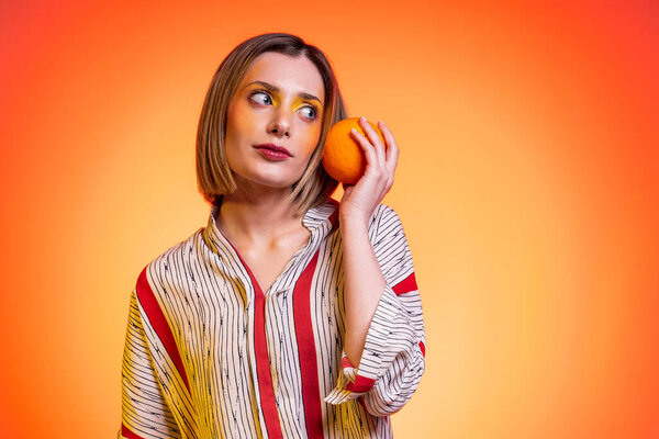 Beautiful attractive blond young woman light blue eyes holding orange as smartphone with bright yellow background - Studio photography of pretty woman who holds citrus in hands like a cell phone