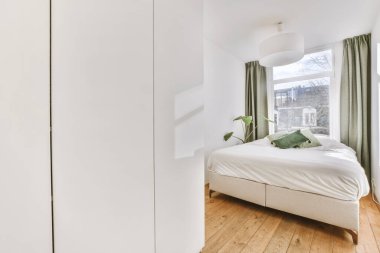 Rahat yatak ve gardırop, modern apartmanın penceresinde beyaz duvarları ve yeşil perdeleri olan dar minimalist yatak odasına yerleştirilmiş.