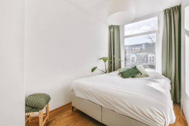 Rahat yatak ve sandalye, modern apartmanın penceresinde beyaz duvarları ve yeşil perdeleri olan dar minimalist yatak odasına yerleştirilmiş.