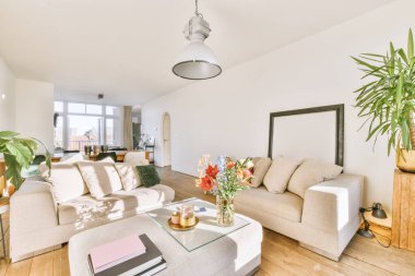 Yumuşak yastıklı rahat koltuklar, lambanın altında vazo olan masa ve geniş ışıklı oturma odasında yemek alanı olan bitki.