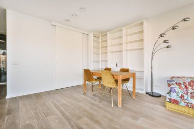 Tahta sandalyeler, masa, duvardaki beyaz raflar boş bir odanın köşesinde.