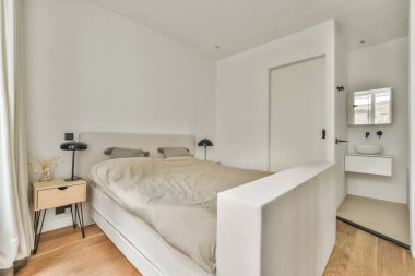 Beyaz çarşaflı rahat bir yatak ve lambasıyla pencere ve balkon kapısının yakınına yerleştirilmiş aydınlık yatak odası.