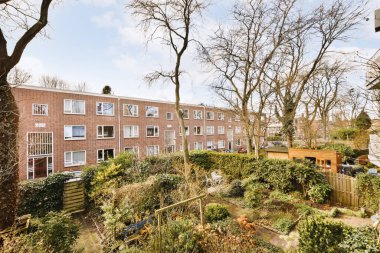 Geniş bir bahçenin panoramik manzarası, ağaçlar, beyaz çerçeveli tuğla evler ve arabalar..