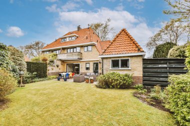 Üç katlı tuğladan bir ev, balkon ve yeşil çimenli geniş bir avlu ve oturma alanı var.