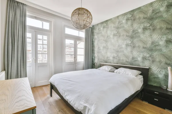 Comfortable bed located near the balcony door with green curtains in a cozy bright bedroom at home