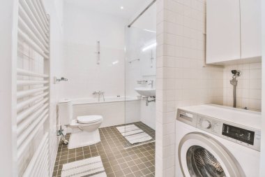 Beyaz banyo ve tuvalet, lavabo yanında cam bölme ve çamaşır makinesi.