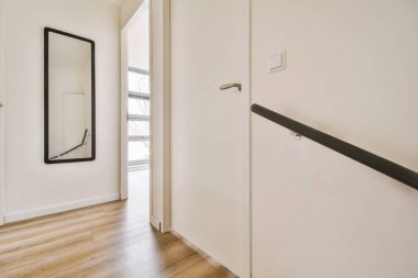 Dar koridor, siyah çerçeveli ayna, beyaz duvarlar ve modern bir dairede pencereleri ve parke döşemeleri olan geniş bir odaya açılan kapılar.