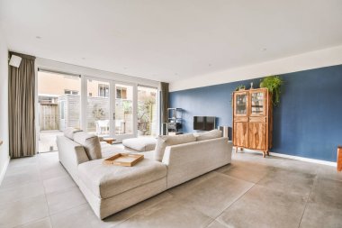 Oturma odasının iç tasarımı modern minimalist tarzda geniş pencereli ve avluya açılan kapısı olan gri bir kanepe, ahşap bir gardırop ve mavi bir duvarda televizyon.