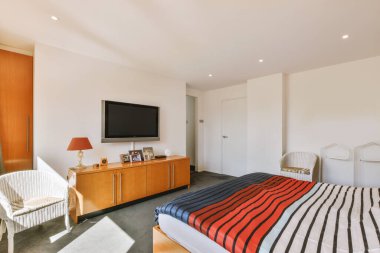 Parlayan lambanın yanında rahat bir yatak ve küçük pencereli ve televizyonlu çağdaş yatak odasında çerçeveli resimler.