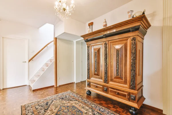 Evin koridorunda açık oturma odası kapısı ve merdivenlerin yanında dekorasyon ve ayna bulunan eski bir dolap.