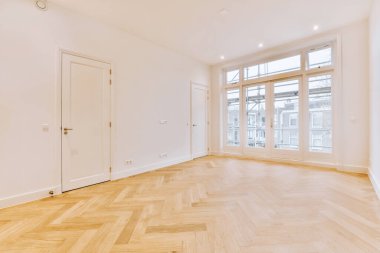 Kapıları, geniş balkon pencereleri ve ahşap parke zemini olan boş beyaz bir oda.