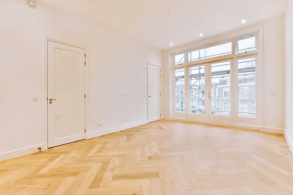 Interior of an empty white room with doors and with large balcony windows and wooden parquet floor