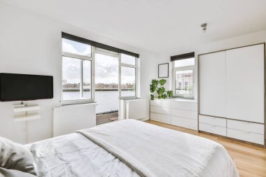 Pencerenin yanında beyaz çarşaflar, balkon kapısı ve karşıdaki aydınlık yatak odasında gardıropları olan rahat bir yatak.