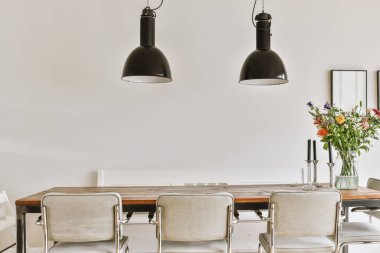 Beyaz duvarlı modern bir oturma odasında, siyah kolye lambalarının altında bir vazo çiçekli ahşap masa.