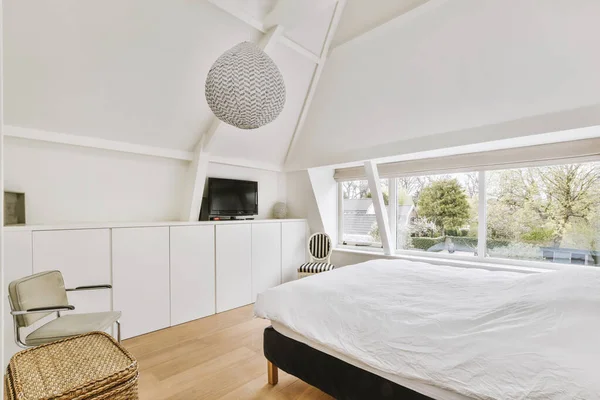 Modern interior design of a small attic bedroom with white walls and a soft cozy bed, storage places, a panoramic window and a TV in front of the bedamic window