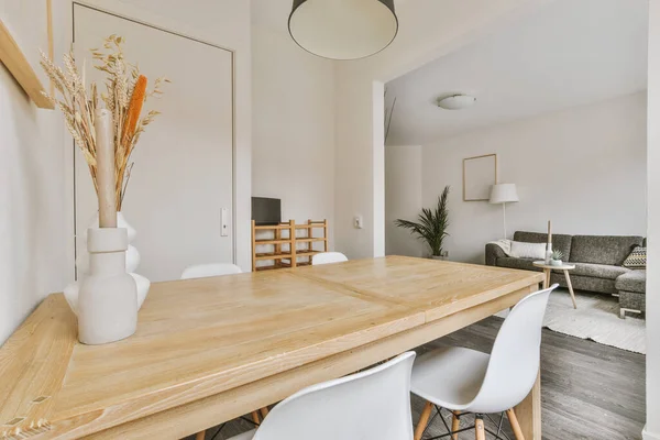 Modern minimalist interior design of a bright apartment with a wooden table and chairs in the dining area next to the living room with white walls and a gray sofa