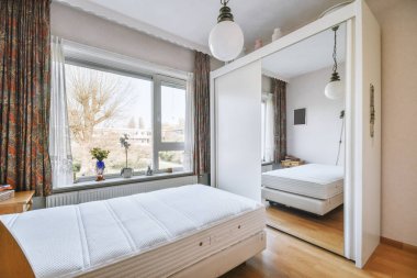 Pencerenin yanındaki cam yatağın yanında rahat bir yatak ve parlak bir yatak odasında aynalı bir gardırop.