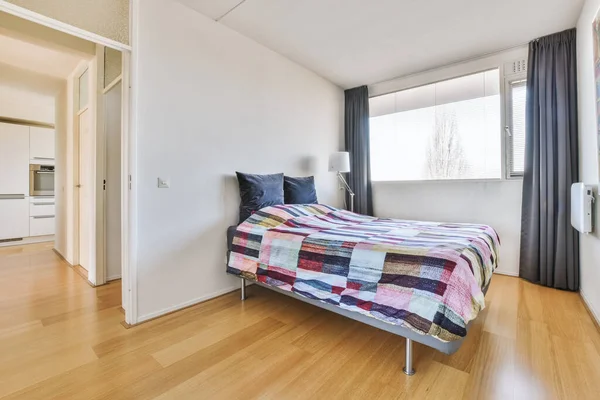 Comfortable bed with a bright blanket and pillows, located by the window with a curtain in the bright bedroom of a modern apartment