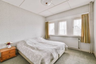 Beyaz çarşaflı rahat bir yatak ve lambasıyla pencere ve balkon kapısının yakınına yerleştirilmiş aydınlık yatak odası.