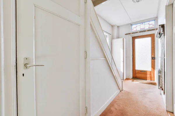 Light hallway with entrance door in two level apartment with minimalist wooden staircase leading upstairs in daylight